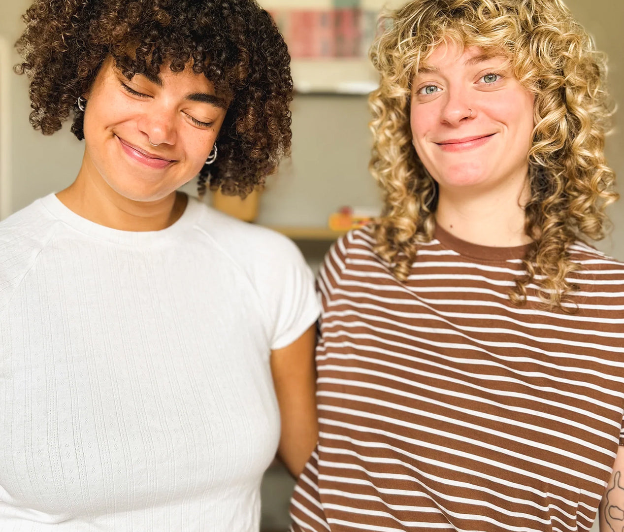 Two smiling women standing indoors, one with dark curly hair wearing a white short-sleeve top and the other with blonde curly hair wearing a brown-and-white striped top.