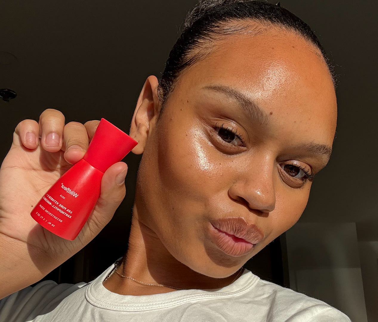 A close-up selfie of a woman holding a small red bottle of Tom Ford Clear Liquid Cosmetics Clear Primer against a dark indoor background.